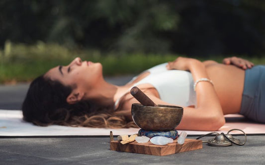 Femme allongée sur un tapis de yoga les yeux fermés, une main sur la poitrine, l'autre sur le ventre ; à côté d'elle un bol tibétain et des pierres minérales.