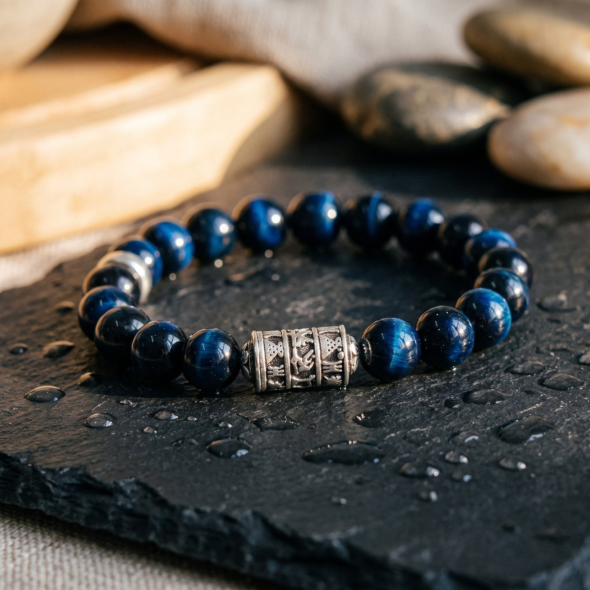Gros plan du bracelet OM en œil de faucon sur une ardoise mouillée, perles bleu nuit chatoyantes et perle tibétaine gravée en argent Image