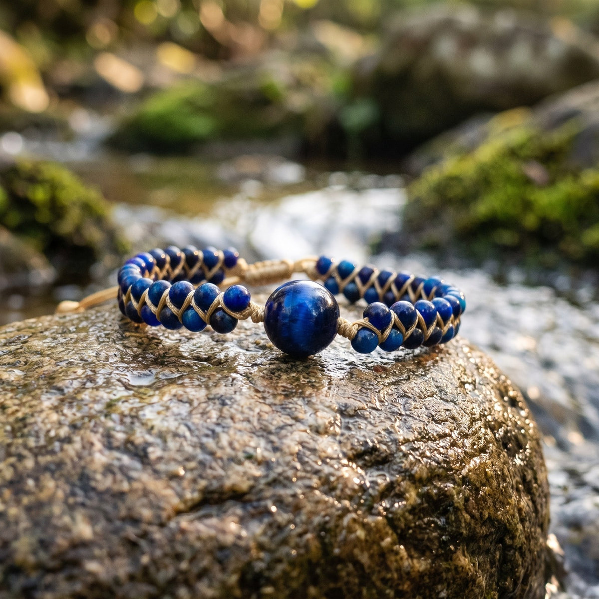 Gros plan du bracelet tressé en œil de faucon posé sur une pierre plate au bord d’un ruisseau, perles bleues et cordon doré Image