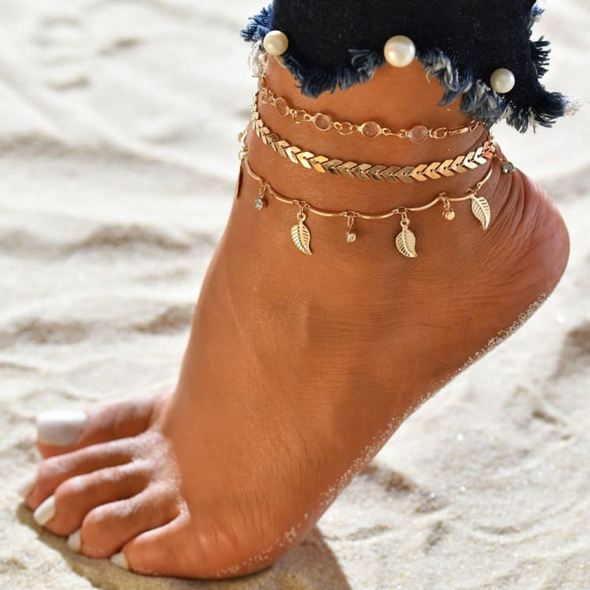 Bracelet de cheville boheme triple rang dore porte a la cheville sur sable, jean en denim perle