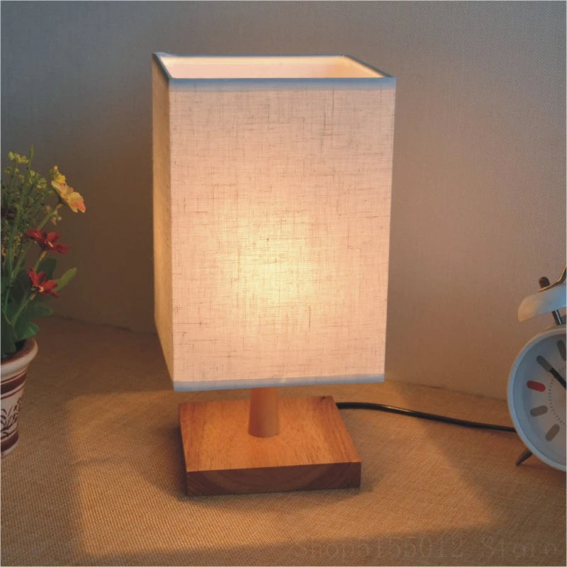Lampe de table zen LED avec socle carré en bois sur fond gris avec une plante à gauche et un réveil à droite